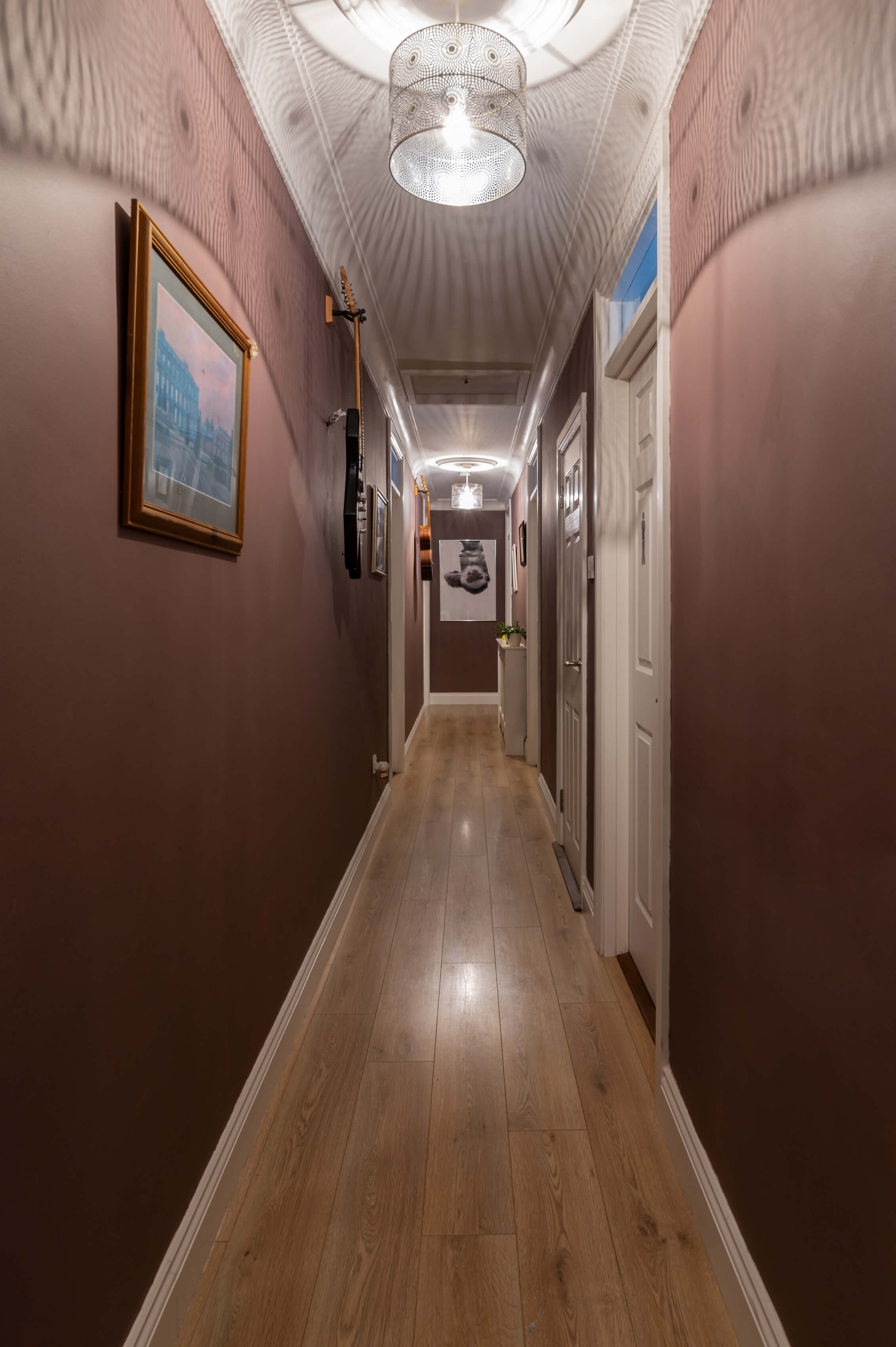 a long bungalow hallway, painted in a dusky purple colour. Decorative laser cut shades on the ceiling and photographs on the walls. Multiple doors along the hallway.